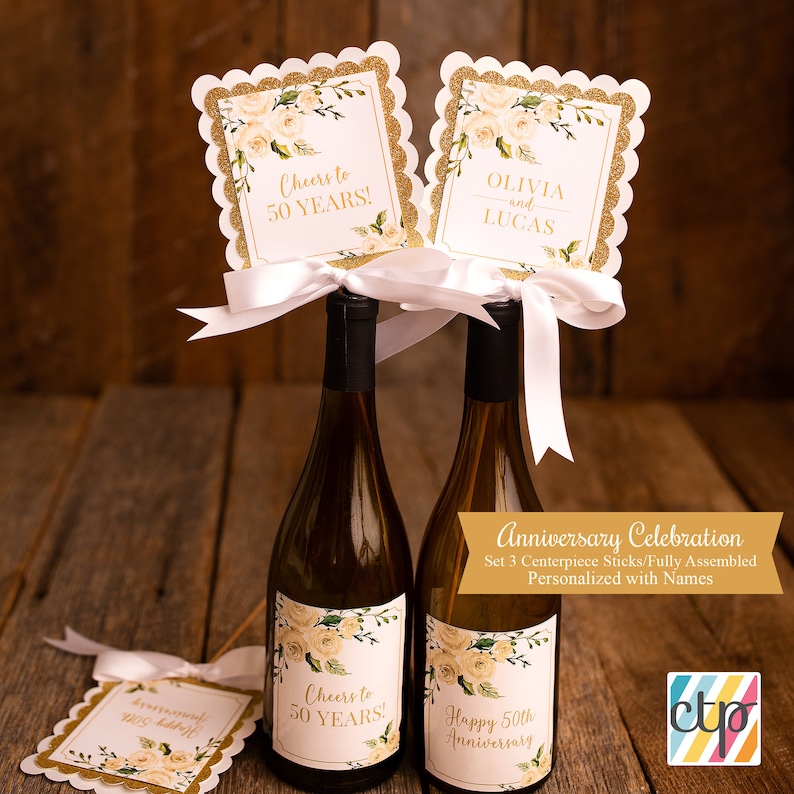 K&ouml;nnte beinhalten: Set aus drei goldenen und wei&szlig;en floralen Flaschenverschl&uuml;ssen mit dem Text "Cheers to 50 Years!" und "Olivia und Lucas". Die Verschl&uuml;sse sind mit einem wei&szlig;en Satinband verziert und bereit, auf Weinflaschen f&uuml;r eine 50. Jubil&auml;umsfeier platziert zu werden.