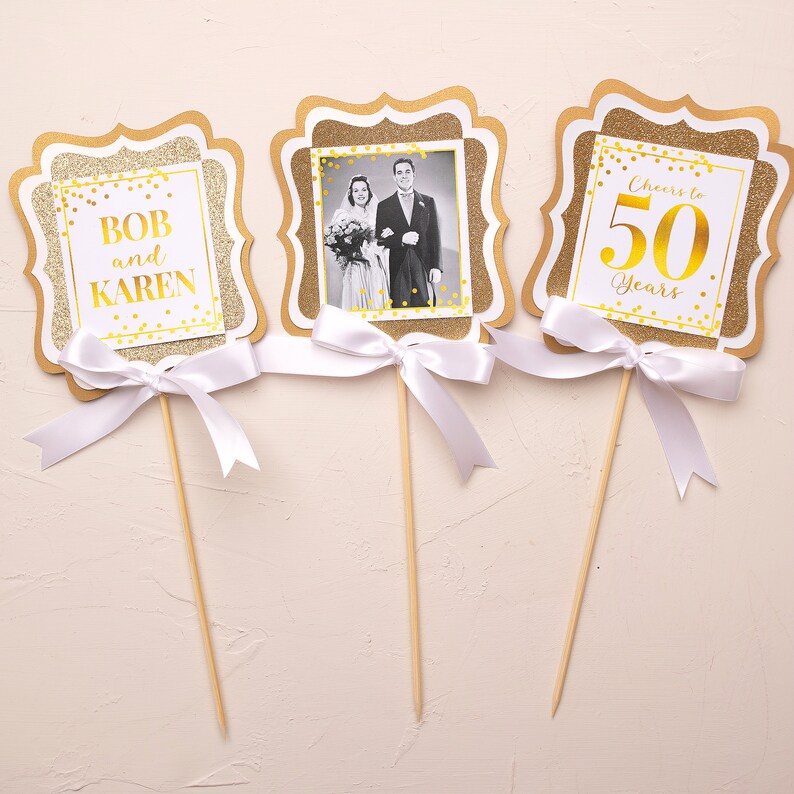 May include: Three gold glitter cake toppers with white ribbon bows. The first topper says "Bob and Karen" in gold lettering. The second topper has a black and white photo of a couple in a frame. The third topper says "Cheers to 50 Years" in gold lettering.