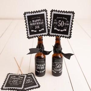 Puede incluir: Cuatro carteles cuadrados negros y plateados con texto blanco. Los carteles dicen "Happy Birthday Joe", "Aged 50 Years", "Vintage Dude 50", y "Cheers to 50 Years". Dos botellas de vidrio marr&oacute;n con cintas negras est&aacute;n en el centro de la imagen.