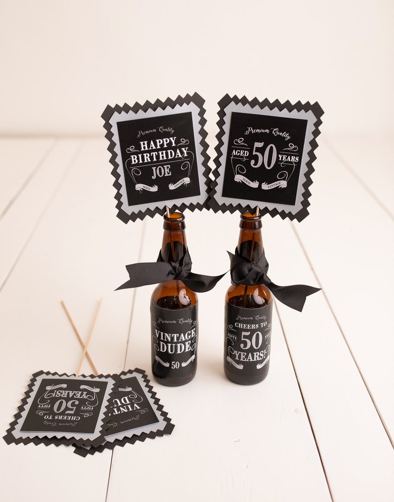 Puede incluir: Cuatro carteles cuadrados negros y plateados con texto blanco. Los carteles dicen "Happy Birthday Joe", "Aged 50 Years", "Vintage Dude 50", y "Cheers to 50 Years". Dos botellas de vidrio marr&oacute;n con cintas negras est&aacute;n en el centro de la imagen.
