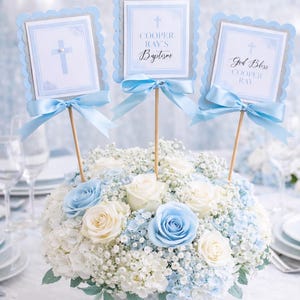 May include: A floral centerpiece with white, light blue, and blue roses, hydrangea, and baby's breath. Three decorative signs on wooden sticks with blue bows read "Cooper Ray's Baptism" and "God Bless Cooper Ray". The arrangement is in a white vase.
