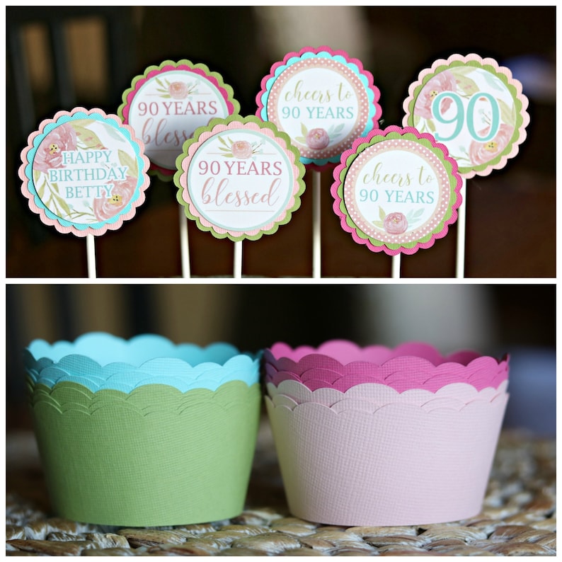 May include: A collection of decorative cupcake toppers and cupcake liners. The toppers feature floral designs and text that reads "Happy Birthday Betty", "90 Years Blessed", and "Cheers to 90 Years". The liners are green, blue, and pink.