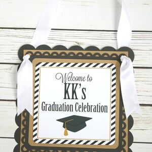 May include: Black and gold graduation party sign with white ribbon. The sign says "Welcome to KK's Graduation Celebration" with a black graduation cap with a gold tassel.