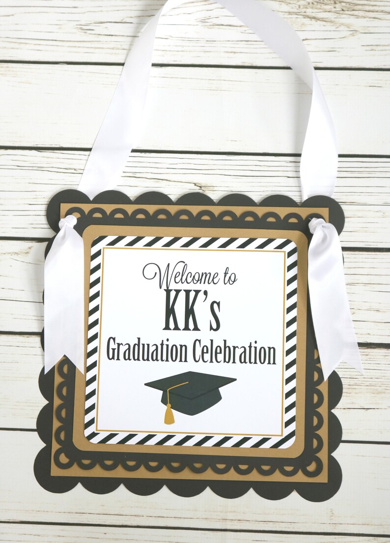 May include: Black and gold graduation party sign with white ribbon. The sign says "Welcome to KK's Graduation Celebration" with a black graduation cap with a gold tassel.