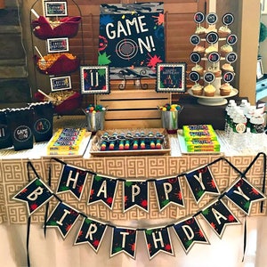 May include: A birthday party table decorated with a 'Game On!' theme. The table is covered in a brown and white tablecloth with a geometric pattern. There are black and green 'Happy Birthday' banners with paint splatters. The table is decorated with black and white signs, candy, and cupcakes. The cupcakes are decorated with black and white frosting and 'Game On!' toppers.