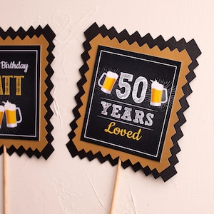 May include: Two black and gold square cake toppers with a scalloped edge. The first topper says "Happy Birthday Matt" with two beer mugs. The second topper says "50 Years Loved" with two beer mugs.