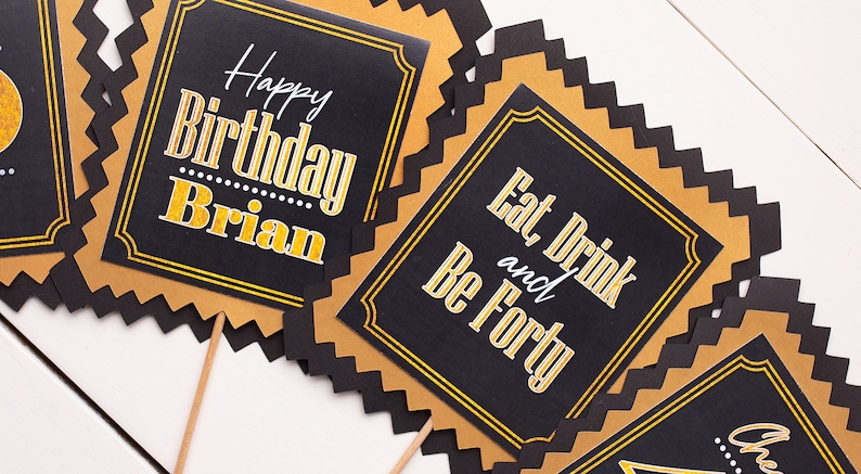 May include: Four black and gold party signs with scalloped edges. The signs have gold lettering on a black background. The text on the signs reads "Happy Birthday Brian", "Eat and Drink", "Be Forty", and "Cheers to 40".