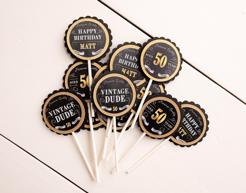 May include: Black and gold cupcake toppers with the text "Happy Birthday Matt", "Vintage Dude", and "Aged 50 Years".