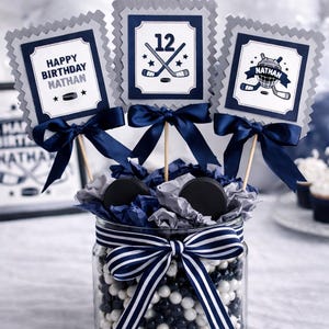 May include: A birthday centerpiece featuring hockey-themed decorations. A glass jar filled with black and white beads is adorned with a navy and white striped bow. Three square toppers with hockey graphics and bows are on wooden sticks.