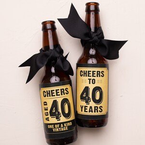 May include: Two brown glass beer bottles with black ribbon bows. The bottles have gold labels with black text that reads "Cheers to 40 Years" and "Aged 40 One of a Kind Vintage".