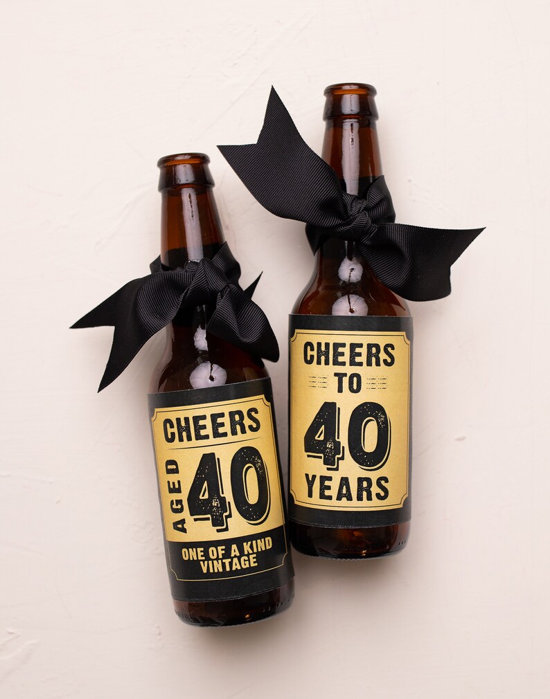 May include: Two brown glass beer bottles with black ribbon bows. The bottles have gold labels with black text that reads "Cheers to 40 Years" and "Aged 40 One of a Kind Vintage".