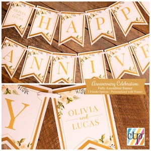 K&ouml;nnte beinhalten: Ein wei&szlig;-goldenes Banner mit den Worten "Happy Anniversary" und den Namen "Olivia und Lucas". Das Banner ist mit wei&szlig;en Rosen und gr&uuml;nen Bl&auml;ttern verziert.