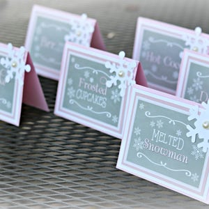 May include: A collection of pink and gray table cards with white snowflake accents. Each card displays a different menu item, such as "Snowflake Cookies" and "Melted Snowman", perfect for a winter-themed event.