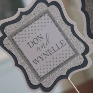 May include: A silver and black glitter card with a white square in the centre. The text "DON and WYNELLE" is printed in black on the white square.