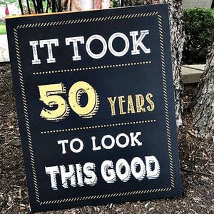 May include: Black chalkboard sign with gold lettering that reads "It took 50 years to look this good".