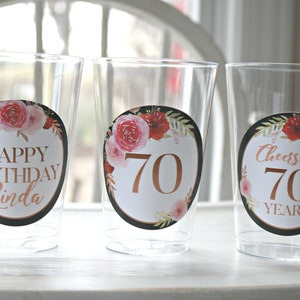 May include: Three clear plastic cups with floral themed stickers. The stickers have a black background with gold lettering. The first cup says "Happy Birthday Linda". The second cup says "70". The third cup says "Cheers to 70 Years".