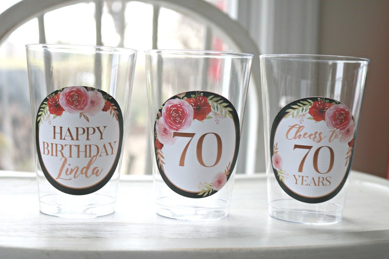 May include: Three clear plastic cups with floral themed stickers. The stickers have a black background with gold lettering. The first cup says "Happy Birthday Linda". The second cup says "70". The third cup says "Cheers to 70 Years".