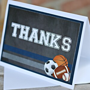 May include: A white card with a blue and gray striped background. The card says "THANKS" in large white letters. There are three sports balls on the card: a basketball, a soccer ball, and a football.