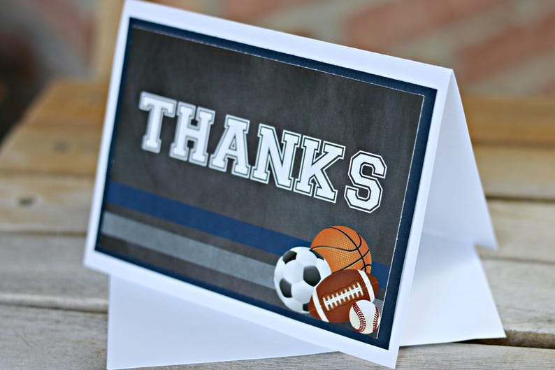 May include: A white card with a blue and gray striped background. The card says "THANKS" in large white letters. There are three sports balls on the card: a basketball, a soccer ball, and a football.
