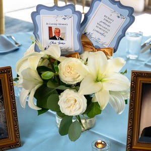 May include: A floral centerpiece with white lilies and roses, accented by greenery, sits on a light blue tablecloth. Two framed cards read "In Loving Memory" and "We won't forget someone who gave us so much to remember."