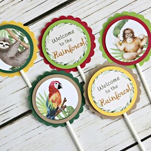 May include: Four cupcake toppers with rainforest animal designs. The toppers feature a sloth, a monkey, a parrot, and a sign that says "Welcome to the Rainforest!"