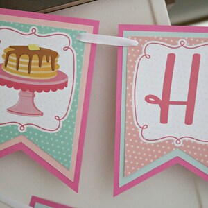 May include: A pink and green banner with the letter "H" and a stack of pancakes on a pink pedestal. The banner has a white polka dot pattern and a pink scalloped border.