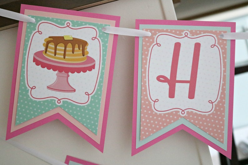 May include: A pink and green banner with the letter "H" and a stack of pancakes on a pink pedestal. The banner has a white polka dot pattern and a pink scalloped border.