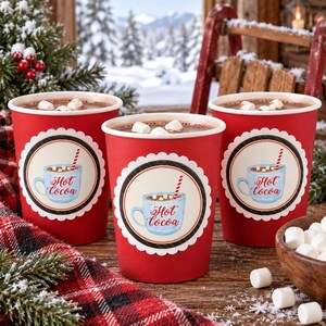 May include: Three red paper cups filled with hot cocoa and marshmallows. Each cup features a circular label with a scalloped white border, a black ring, and an illustration of a light blue mug with the text Hot cocoa and a red striped straw.
