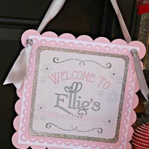 May include: A square decorative sign with scalloped pink edges and a white ribbon hanger. The center features silver glitter borders, snowflake accents, and the text "WELCOME TO Ellie's Winter ONEderland" in pink and silver lettering.