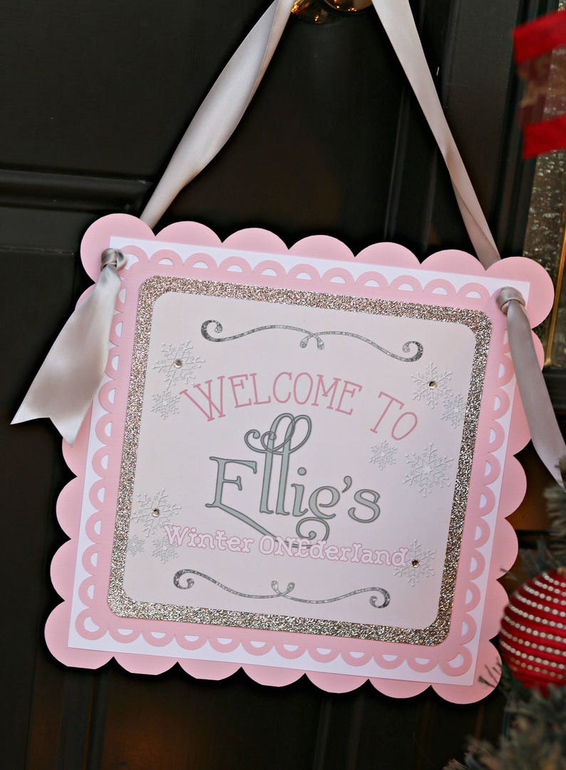May include: A square decorative sign with scalloped pink edges and a white ribbon hanger. The center features silver glitter borders, snowflake accents, and the text "WELCOME TO Ellie's Winter ONEderland" in pink and silver lettering.