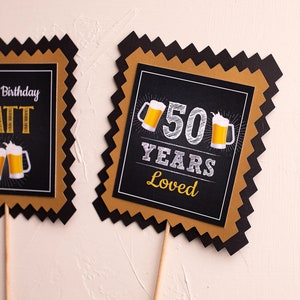 May include: Two black and gold square cake toppers with a scalloped edge. The first topper says "Happy Birthday Matt" with two beer mugs. The second topper says "50 Years Loved" with two beer mugs.