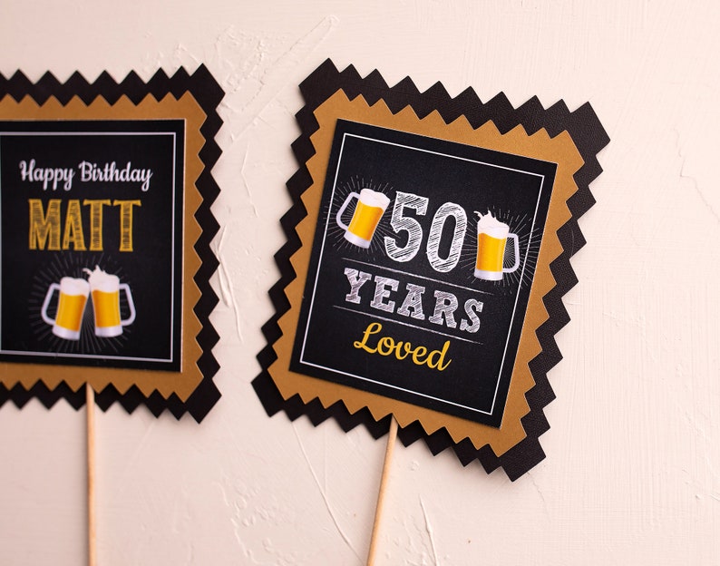 May include: Two black and gold square cake toppers with a scalloped edge. The first topper says "Happy Birthday Matt" with two beer mugs. The second topper says "50 Years Loved" with two beer mugs.