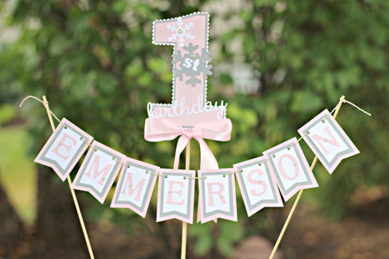 May include: Pink and gray cake topper with a number one, snowflakes, and the text "1st birthday". A pink and gray banner with the name "EMERSON" is attached to the topper.