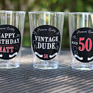 May include: Three clear plastic cups with black and white circular labels. The labels have white text on a black background. The text on the labels reads "Premium Quality Happy Birthday Matt", "Premium Quality Vintage Dude 50", and "Premium Quality Aged 50 Years".
