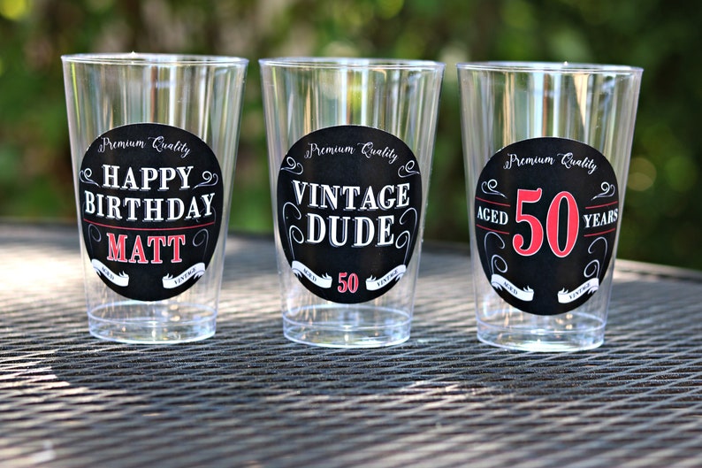 May include: Three clear plastic cups with black and white circular labels. The labels have white text on a black background. The text on the labels reads "Premium Quality Happy Birthday Matt", "Premium Quality Vintage Dude 50", and "Premium Quality Aged 50 Years".