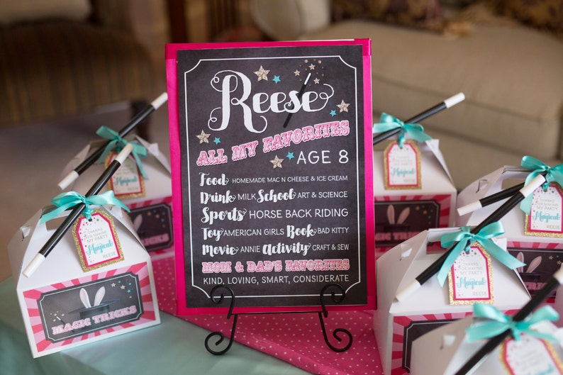 May include: A pink and black chalkboard sign with the text "Reese All My Favorites * Age 8" and a list of Reese's favorite things, including food, drinks, sports, toys, movies, and activities. The sign is on a black stand and there are several white boxes with pink and blue decorations and magic wands on a pink and white table.