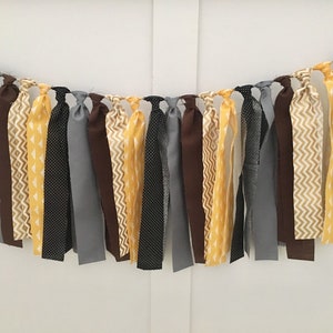 May include: A fabric banner with a mix of yellow, brown, gray, and black fabric strips. The strips are tied together to create a garland.