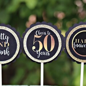 Puede incluir: Tres toppers redondos para cupcakes negros y dorados con bordes festoneados. Los toppers muestran el texto "Betty y Frank", "Cheers to 50 Years" y "Happy Anniversary".