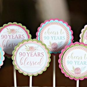 May include: A collection of decorative cupcake toppers for a 90th birthday celebration. Each topper features a scalloped edge in pink, green, and blue, with floral designs and text such as "Happy Birthday Betty" and "90 Years Blessed."