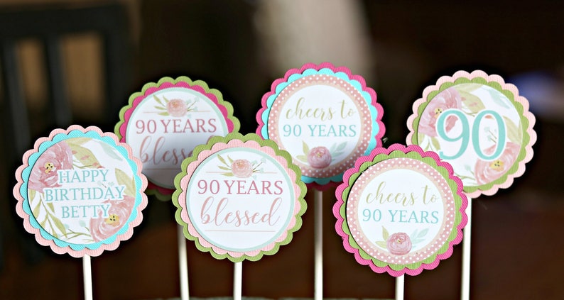 May include: A collection of decorative cupcake toppers for a 90th birthday celebration. Each topper features a scalloped edge in pink, green, and blue, with floral designs and text such as "Happy Birthday Betty" and "90 Years Blessed."