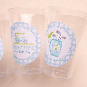 May include: Transparent plastic cups with golf-themed stickers. One sticker shows a golf cart, the text "ANTHONY'S first birthday", and a blue gingham border. Another sticker shows golf clubs in a bag with the number "1" and a green flag.
