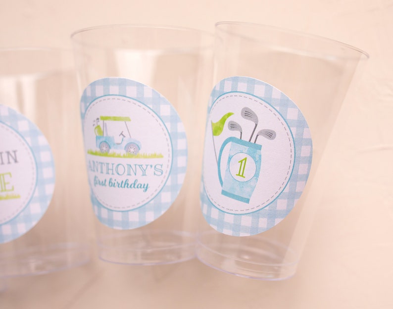May include: Transparent plastic cups with golf-themed stickers. One sticker shows a golf cart, the text "ANTHONY'S first birthday", and a blue gingham border. Another sticker shows golf clubs in a bag with the number "1" and a green flag.