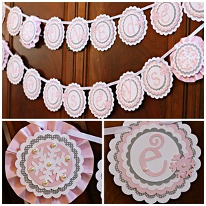 May include: A pink and gray banner with the word "One" spelled out in white letters. Each letter is on a separate circle with a scalloped edge. The circles are connected by a white ribbon. The banner is decorated with snowflakes and pearls.