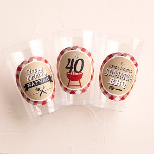May include: Three clear plastic cups with red and white checkered paper labels. The labels have a grill and barbecue theme. One cup says "Happy Birthday Nathan" with a grill and spatula. Another cup says "Grill & Chill Summer BBQ" with a grill and spatula. The third cup says "40" with a grill and flames.