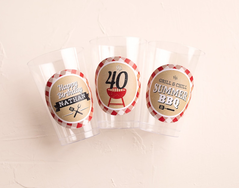 May include: Three clear plastic cups with red and white checkered paper labels. The labels have a grill and barbecue theme. One cup says "Happy Birthday Nathan" with a grill and spatula. Another cup says "Grill & Chill Summer BBQ" with a grill and spatula. The third cup says "40" with a grill and flames.