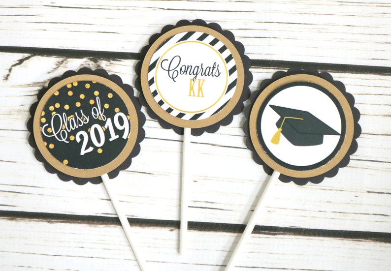 May include: Three black and gold graduation cupcake toppers. One topper has gold glitter dots and says "Class of 2019". Another topper has black and white stripes and says "Congrats KK". The last topper has a black graduation cap with a gold tassel.