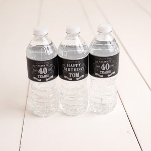 Puede incluir: Tres botellas de agua de pl&aacute;stico transparente con etiquetas negras y plateadas. Las etiquetas dicen "Cheers to 40 Years!" y "Happy Birthday Tom".