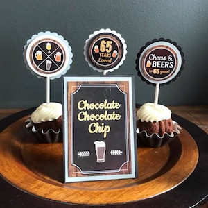 May include: A wooden platter with two chocolate cupcakes topped with white frosting and a sign that reads "Chocolate Chocolate Chip". There are three cupcake toppers with beer-themed designs, one that says "65 Years Loved", and one that says "Cheers & Beers to 65 Years!"