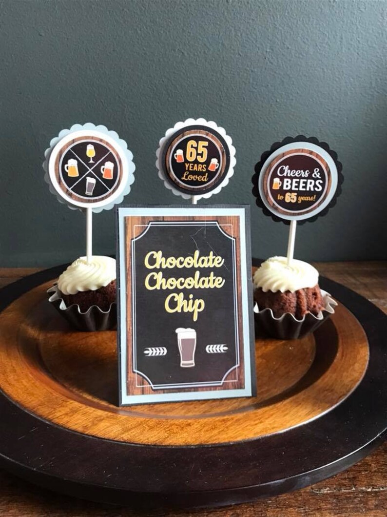 May include: A wooden platter with two chocolate cupcakes topped with white frosting and a sign that reads "Chocolate Chocolate Chip". There are three cupcake toppers with beer-themed designs, one that says "65 Years Loved", and one that says "Cheers & Beers to 65 Years!"