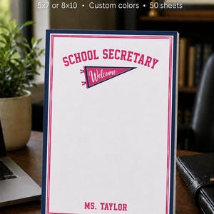May include: A white notepad with a dark blue and pink border. The text reads SCHOOL SECRETARY at the top, with a pennant graphic containing the word Welcome in the center, and the name MS. TAYLOR printed in pink at the bottom of the page.